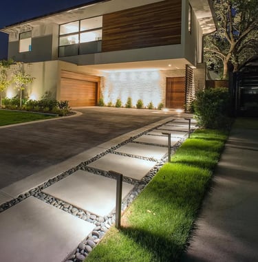 Modern luxury home exterior with illuminated concrete paver walkway and landscape lighting at night.