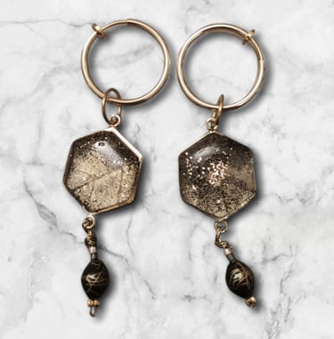 Handmade gold hexagon drop earrings with glitter resin and black beads on a white marble background.
