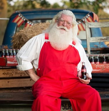 A vintage Santa Claus in red overalls holding a Coca-Cola bottle while sitting on a classic blue truck.