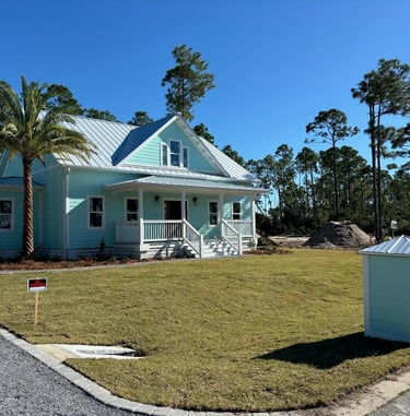 New light blue coastal home with a metal roof, front porch, and palm tree on a grassy lawn.