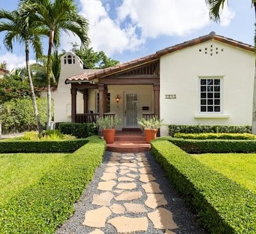 Spanish style bungalow house with a stone walkway, manicured green lawn, and tropical palm trees.