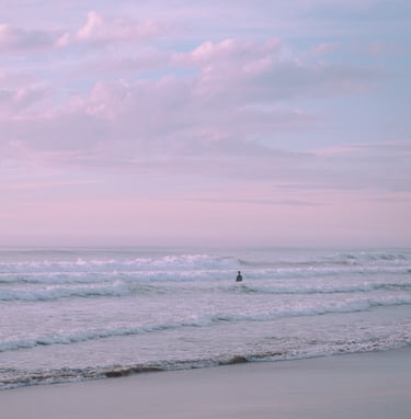 Serene shot of man in the distance standing in the ocean's waves. Pastel sunset sky.