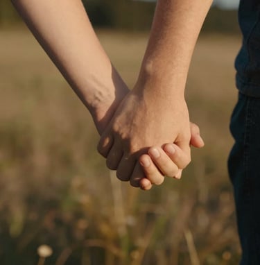 A close-up candid of a child's hand holding a parent's hand, softly lit by the warm afternoon sun. The background is a soft-focus meadow of earthy warm brown and deep charcoal shadows.