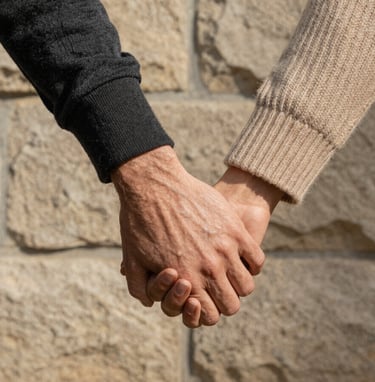 A refined, close-up shot of two people holding hands against a soft sand colored stone wall. One person wears a charcoal sleeve, the other a warm beige knit. The lighting is natural and directional, highlighting the textures and the authentic connection. Elegant and storytelling-focused.