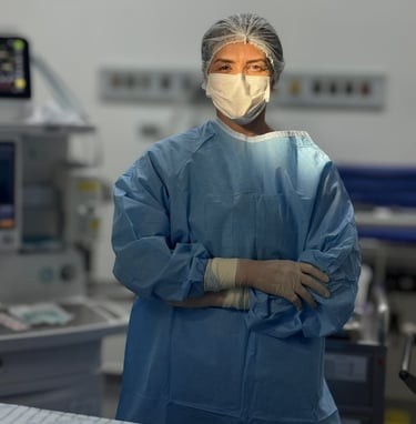 mulher branca em sala de cirurgia paramentada de braços cruzados olhando na direção da camera com um foco de luz em seu rosto