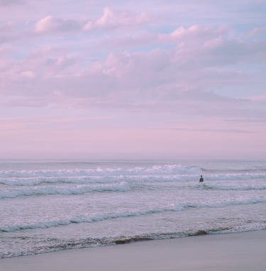 Serene shot of man in the distance standing in the ocean's waves. Pastel sunset sky.