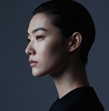 A high-fashion editorial portrait featuring a model in a sharp silhouette. The composition is minimalist, with soft blue-grey lighting hitting the contours of the face against a dark charcoal backdrop. Refined and elegant style.