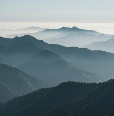 A professional wide-angle landscape photograph of a misty mountain range in the North American Pacific Northwest. The scene is dominated by layers of peaks in dark teal and muted blue-grey, shrouded in soft fog. The composition is minimalist and modern, emphasizing the vast, calm beauty of the wilderness.