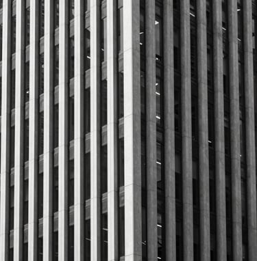 A vertical architectural photograph of a modern building facade featuring clean vertical lines and glass. The image is strictly monochrome with deep blacks and bright whites. Sharp shadows create a geometric pattern. North American / European city center.