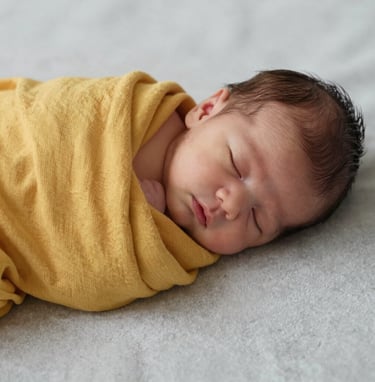 A close-up, high-resolution photo of a sleeping newborn baby wrapped in a soft mustard yellow muslin cloth. The baby is placed on a light grey textured surface. The lighting is soft and natural, suggesting a peaceful Middle Eastern / Turkish nursery setting.