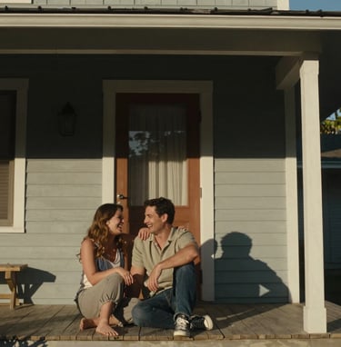 A couple sharing a private, authentic laugh while sitting on the porch of a classic North American / US home. The scene is shot in a cinematic style with warm, sun-drenched lighting and deep charcoal shadows for contrast.