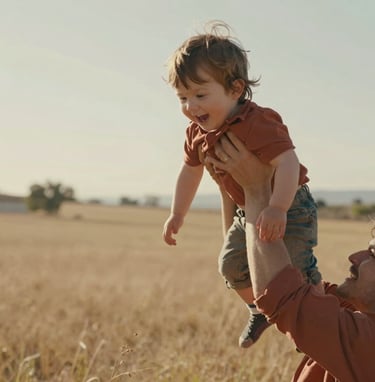 A candid, cinematic close-up of a young child being lifted into the air by a parent in a sun-drenched North American / US meadow. The lighting is warm and hazy, with terracotta and soft sand tones in the clothing and surroundings, highlighting an authentic moment of joy.