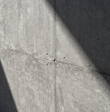 Architectural detail shot of a textured concrete wall with precise joints, showing tectonic rigor. Soft natural light, shadows in dark charcoal grey, material in light grey.