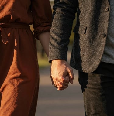 A close-up lifestyle shot of a couple's hands held together during a walk in a North American / US park at golden hour. Warm cinematic lighting with charcoal and terracotta clothing accents.