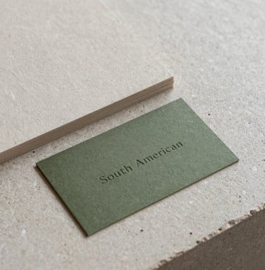 Close-up photography of high-quality tactile paper samples and a minimalist olive green business card on a light gray stone surface, soft natural lighting, South American design studio setting.