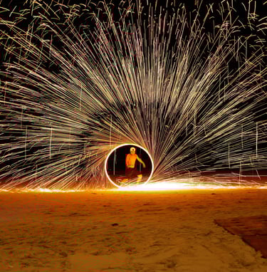 A fire spinner performs on a beach at night, creating a circle of sparks.- by ACAT PHOTOS