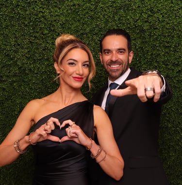 A couple in formal attire posing against a green hedge wall, making a heart symbol and pointing.