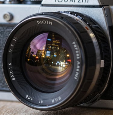 A close-up of a vintage 35mm film camera lens reflecting the lights of a North American / US city at night. The image has a polished, high-end feel with steel gray metallic textures and deep onyx shadows.