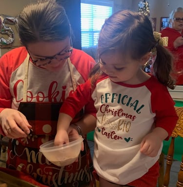 Little girl baking cookies with her aunt wearing a shirt reading, “Official Christmas Cookie Taster”