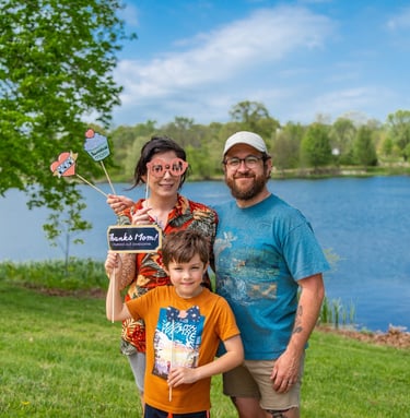 Staged photo of a couple with a young child in front of a lake, holding up paper props