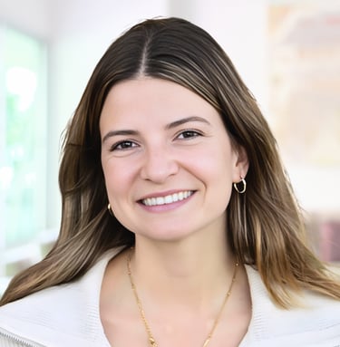 A smiling professional woman with brown hair and gold jewelry wearing a white blazer in an office.