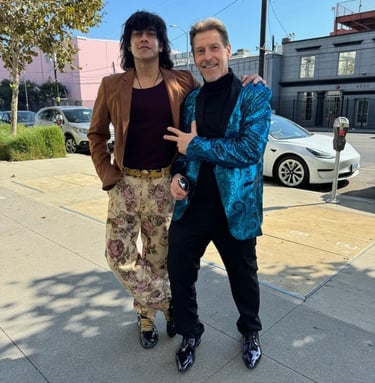 Tim and Bollywood actor in fashionable blazers posing on a sunny urban sidewalk with city buildings.