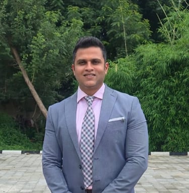 Professional businessman in a grey suit and pink shirt standing outdoors with a green tree background.