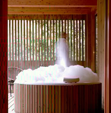 A rustic round cedar hot tub filled with white bubbles on a wooden patio with slatted privacy walls.