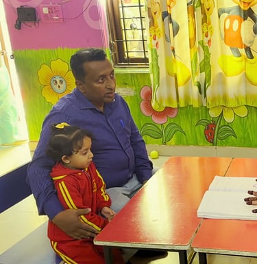 Father and young daughter meeting with a preschool teacher at a small table in a colorful classroom.