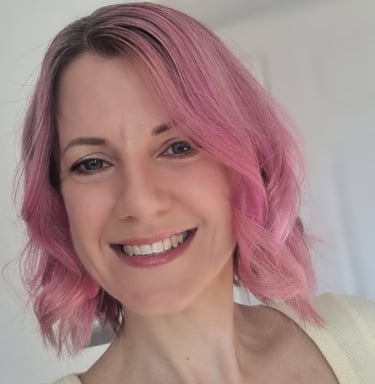 A head and shoulders image of a woman with pink hair, smiling, wearing a yellow top.