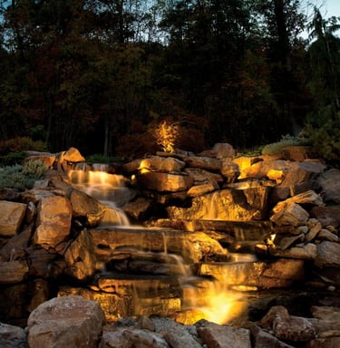 Illuminated backyard stone waterfall feature with landscape lighting and woodland backdrop.