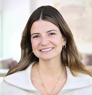 A smiling young woman with long brown hair and a white sweater poses in front of a blurred living room, showcasing a