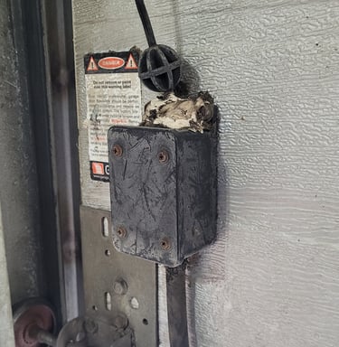 Damaged garage door safety sensor box with exposed wiring and a danger warning label.