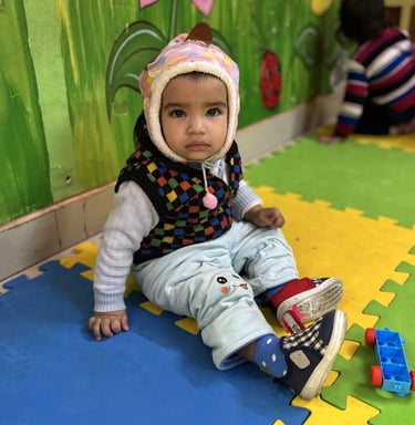 A toddler wearing a warm winter hat sits on colorful foam play mats in a vibrant daycare setting.