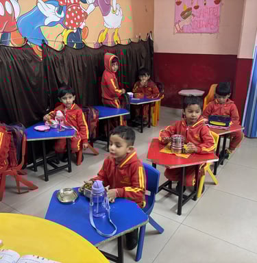 Preschool children in red uniforms sitting at colorful desks during lunch in a classroom with cartoon murals.