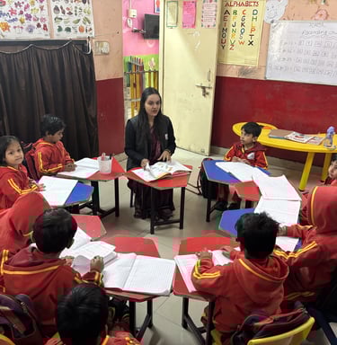 A primary school teacher sits with students in a circle for a lesson in a colorful classroom.