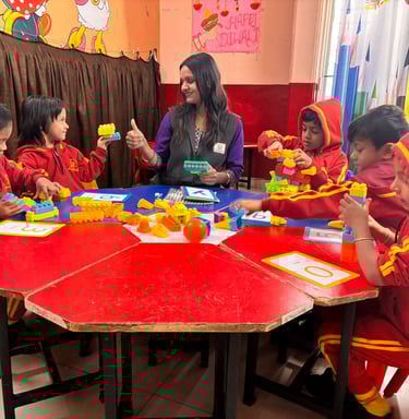 Preschool children in red uniforms building with colorful toy blocks around a table with their teacher.