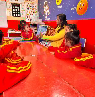 A teacher reads a storybook to preschool students at a red table in a colorful space-themed classroom.