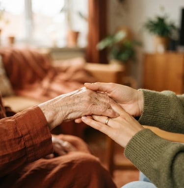Close-up of an elderly person's hand gently held by a younger hand, warm natural window light, shallow depth of field, rich earthy terracotta tones, 35mm.