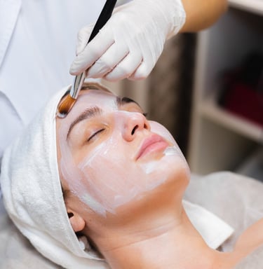 Beautician with a brush applies a white moisturizing mask to the face of a young girl client in a sp