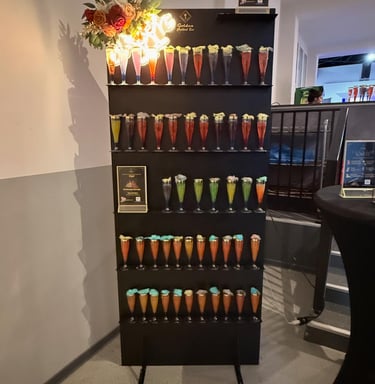 A tiered cocktail display wall at a bar featuring rows of colorful welcome drinks for a party or event.
