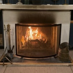 A cozy wood burning fireplace with a mesh screen, iron tools, and glowing embers inside a home.