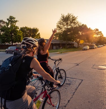 Image of two women bikers riding in the street, one with her arm stretched up in celebration
