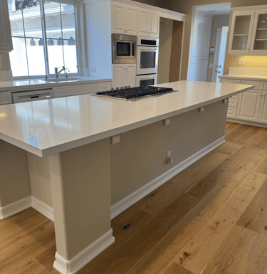 View of a kitchen. There is an island with burners. The background has a sink, microwave, and ovens