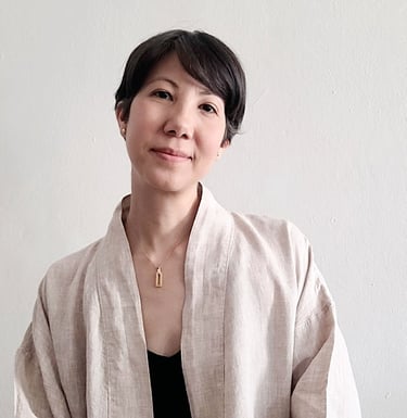 A smiling woman with short black hair wearing a beige linen cardigan and gold necklace against a white wall.