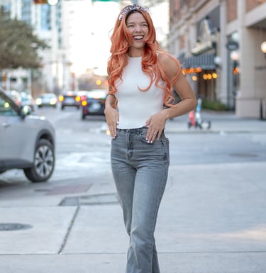 Smiling woman with orange hair wearing a birthday headband and gray jeans on an austin sidewalk.