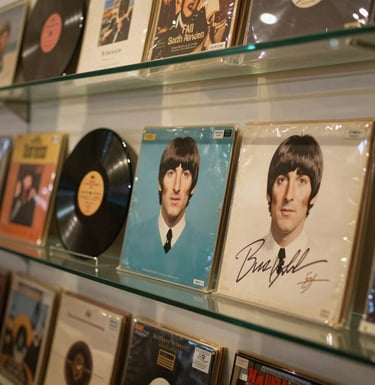 A close-up photograph of a rare Beatles record collection and signed memorabilia displayed behind polished glass in a South American gallery, with warm golden and muted teal tones.