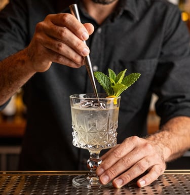 Close-up photography of a mixologist's hands preparing a craft cocktail in a Brazilian bar, focus on the crystal glassware and fresh garnish, warm lighting, charcoal and gold tones in the background.
