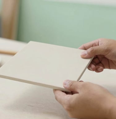 A close-up of a craftsman's hands carefully inspecting a large, matte-finish ceramic panel. The environment is a clean, modern studio with soft green accents #A4BC9F in the background.
