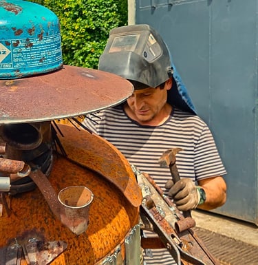 Murat wearing a welding mask and gloves uses a hammer to shape a rusty scrap metal sculpture.